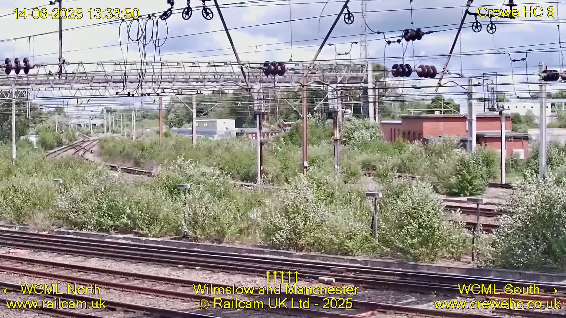 A view of railway tracks and infrastructure overgrown with vegetation under a partly cloudy sky.