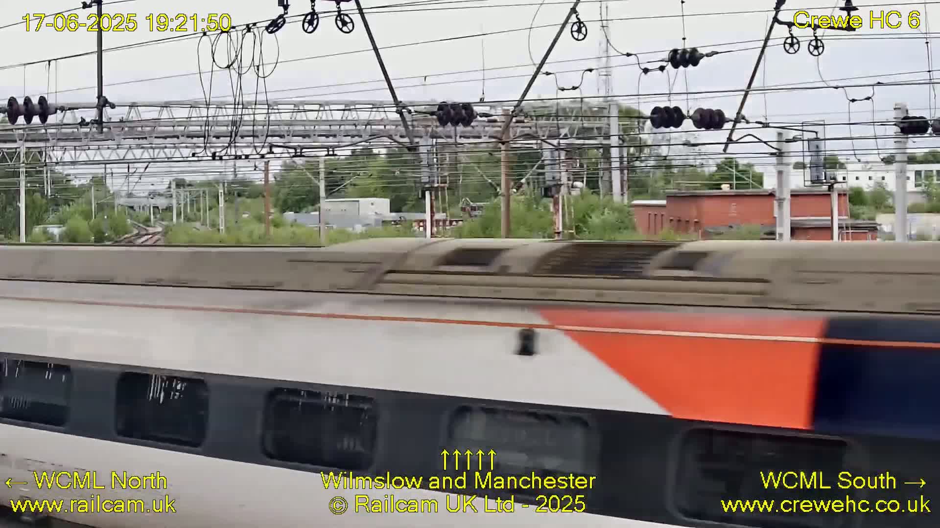 A high-speed train passes by under an overcast sky, beside railway infrastructure and greenery.