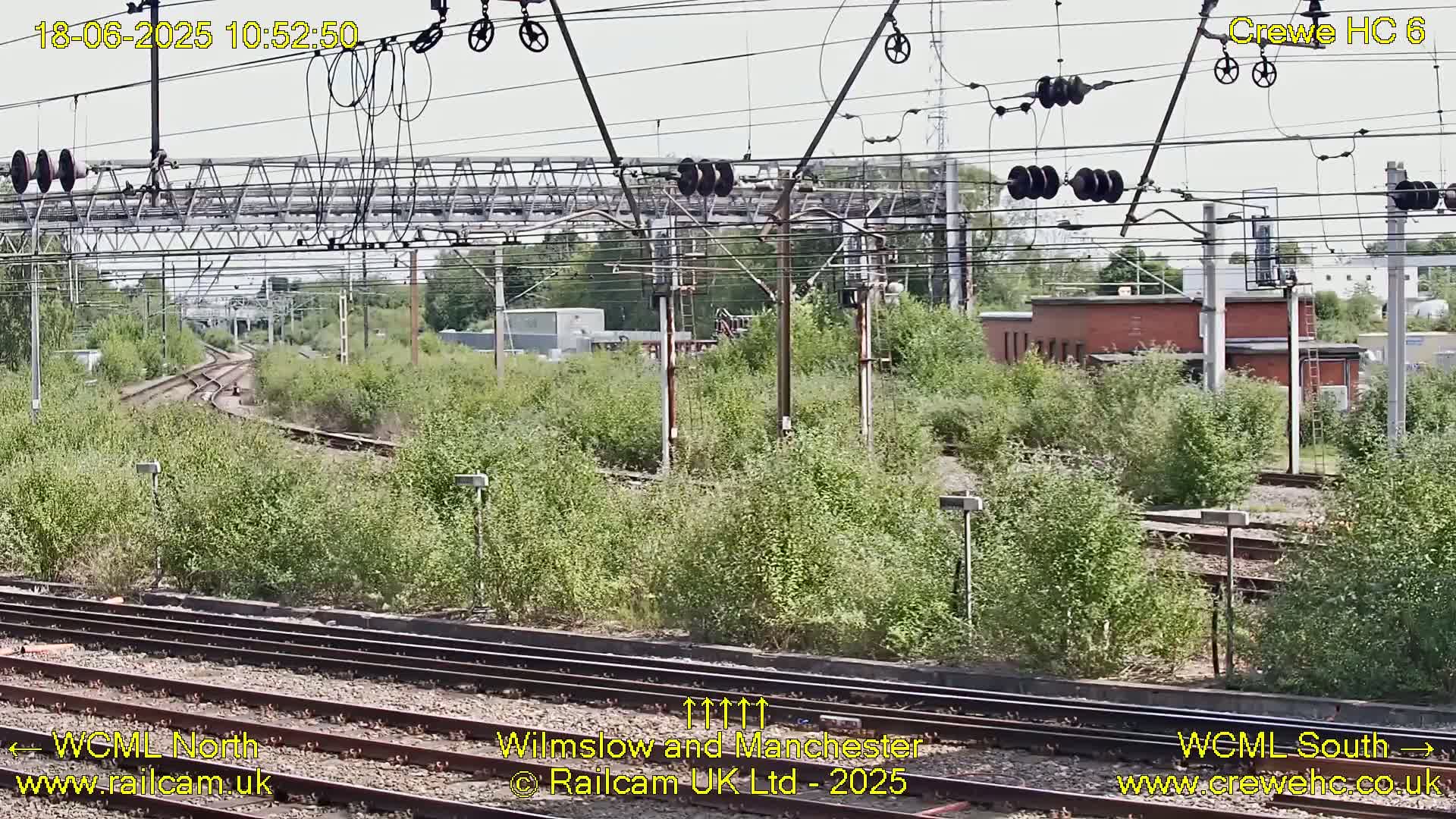 A view of railway tracks and overhead lines on a partly sunny day, with lush green vegetation growing alongside the tracks.
