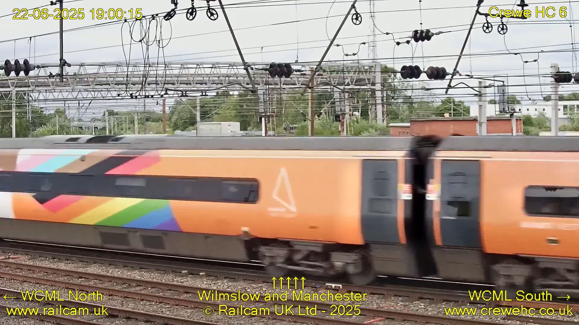 A multicolored, primarily orange train moves past overhead power lines on a cloudy day.