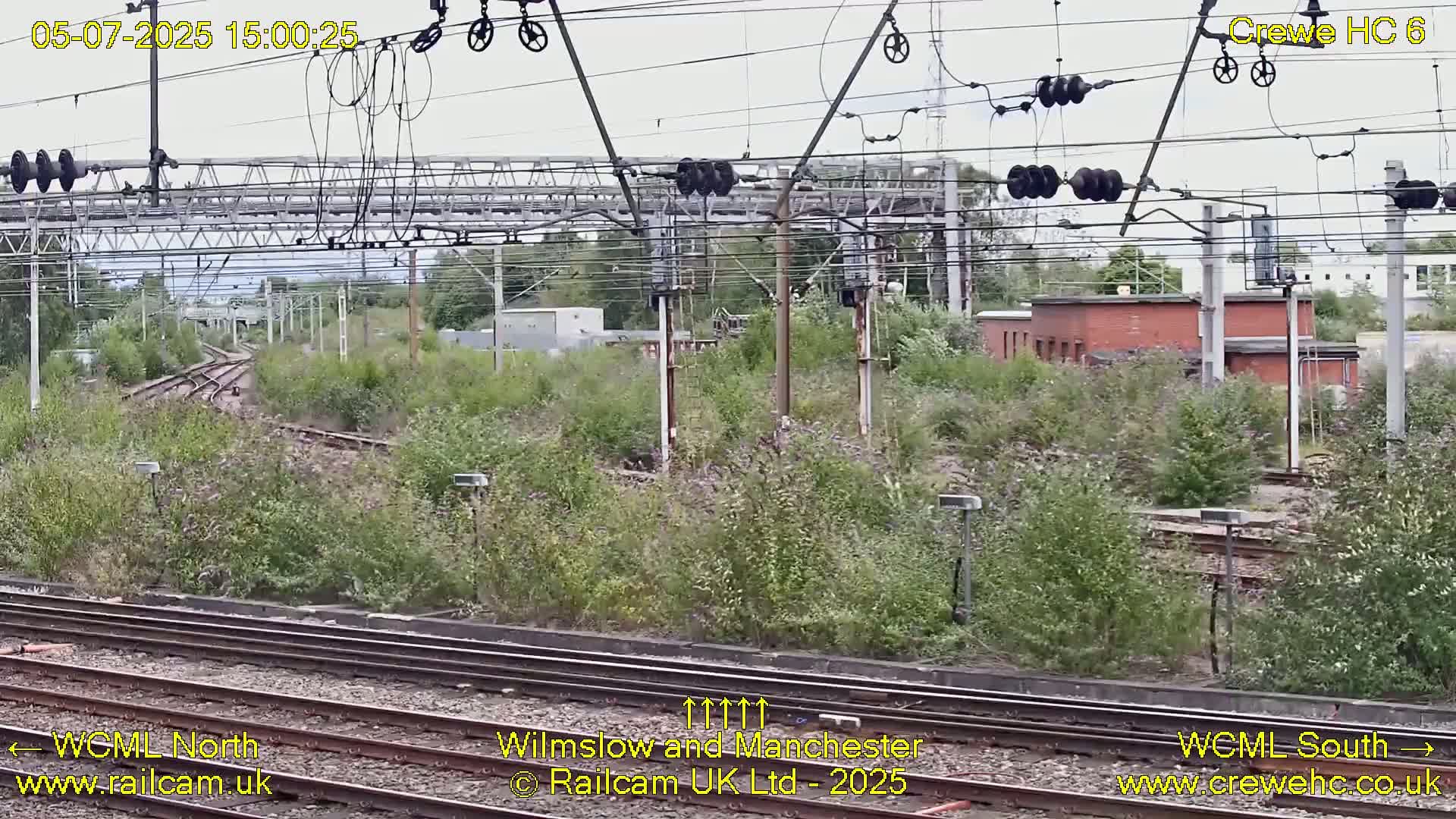 A view of railway tracks and infrastructure overgrown with vegetation under a partly cloudy sky.