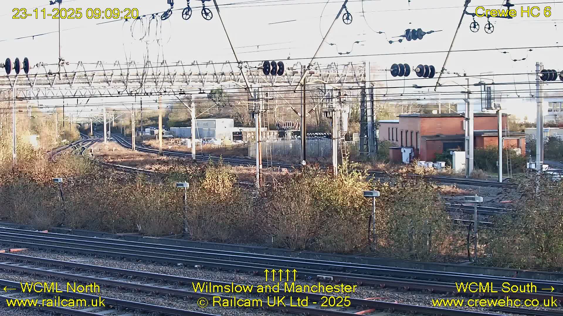 An outdoor daytime scene depicts a complex network of multiple railway tracks with extensive overhead power lines, flanked by dense autumnal bushes and various industrial buildings, all under a bright, clear sky.