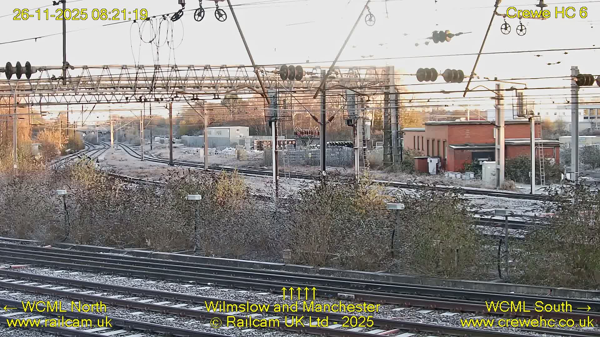 Numerous railway tracks with extensive overhead electrification and industrial buildings in the distance are visible, all lightly covered in frost on a chilly, bright morning.