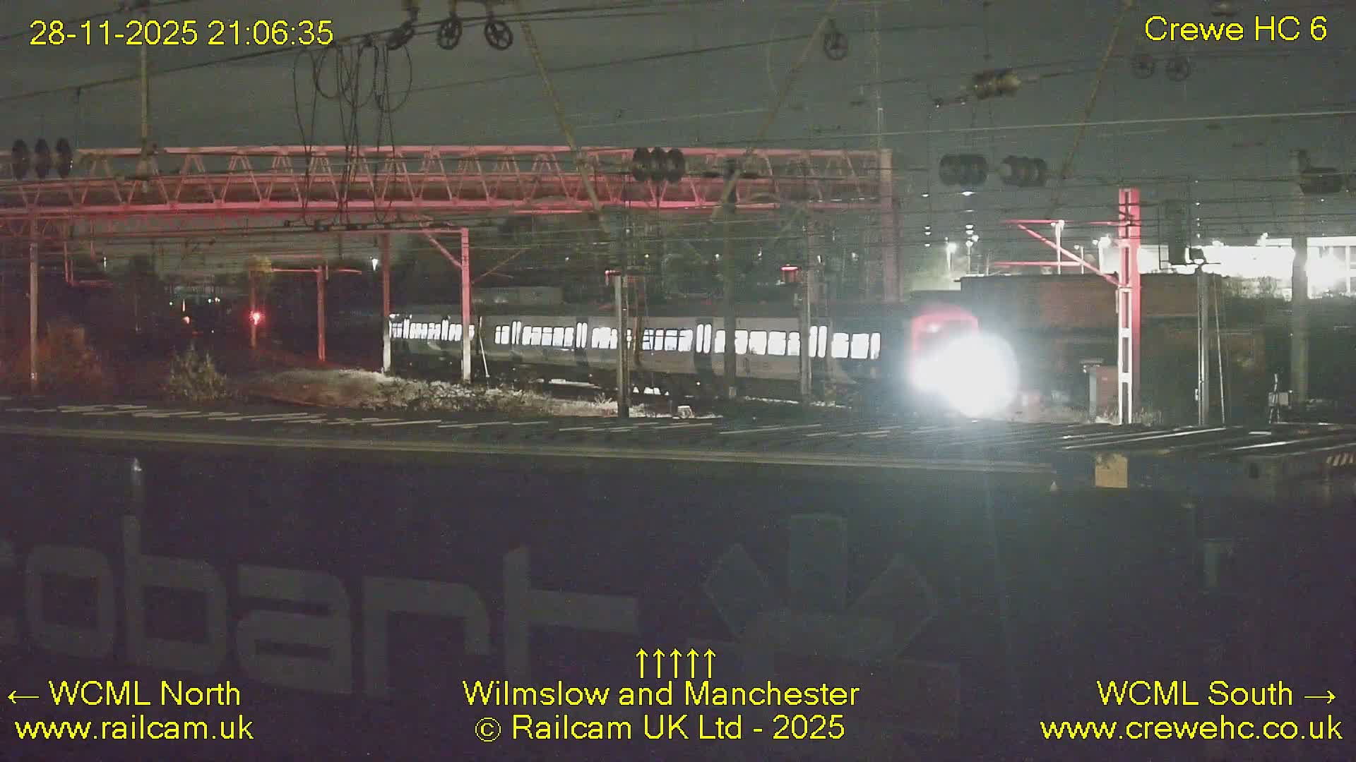At night, a bright-headlighted train approaches on the right while another train with illuminated windows is visible on the left, set against a backdrop of railway tracks and overhead gantries under clear skies.