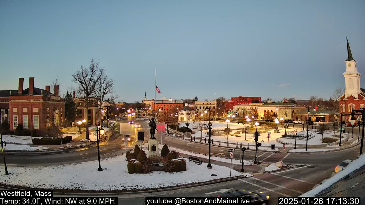 Here's a description of the image: A snow-dusted town square at twilight, with brick buildings, streetlights, and a statue in the center, under a clear, calm sky.