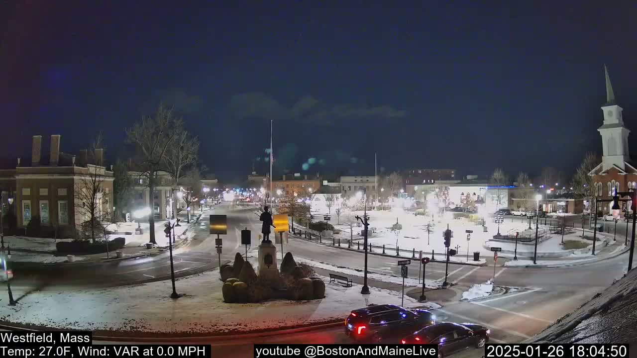 A snow-covered town square at night is illuminated by streetlights, with a few cars visible and a statue in the center.