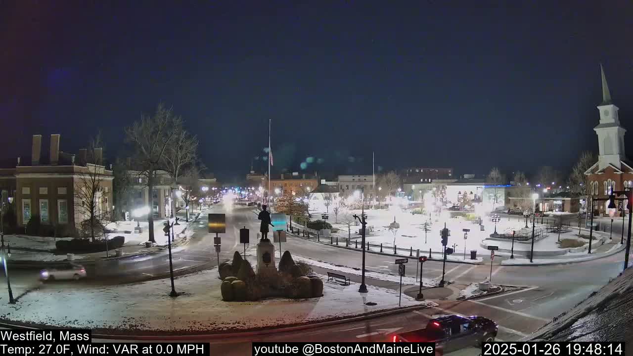 A snow-covered town square at night, illuminated by streetlights, features a statue in the center and a church in the background under a clear, dark sky.