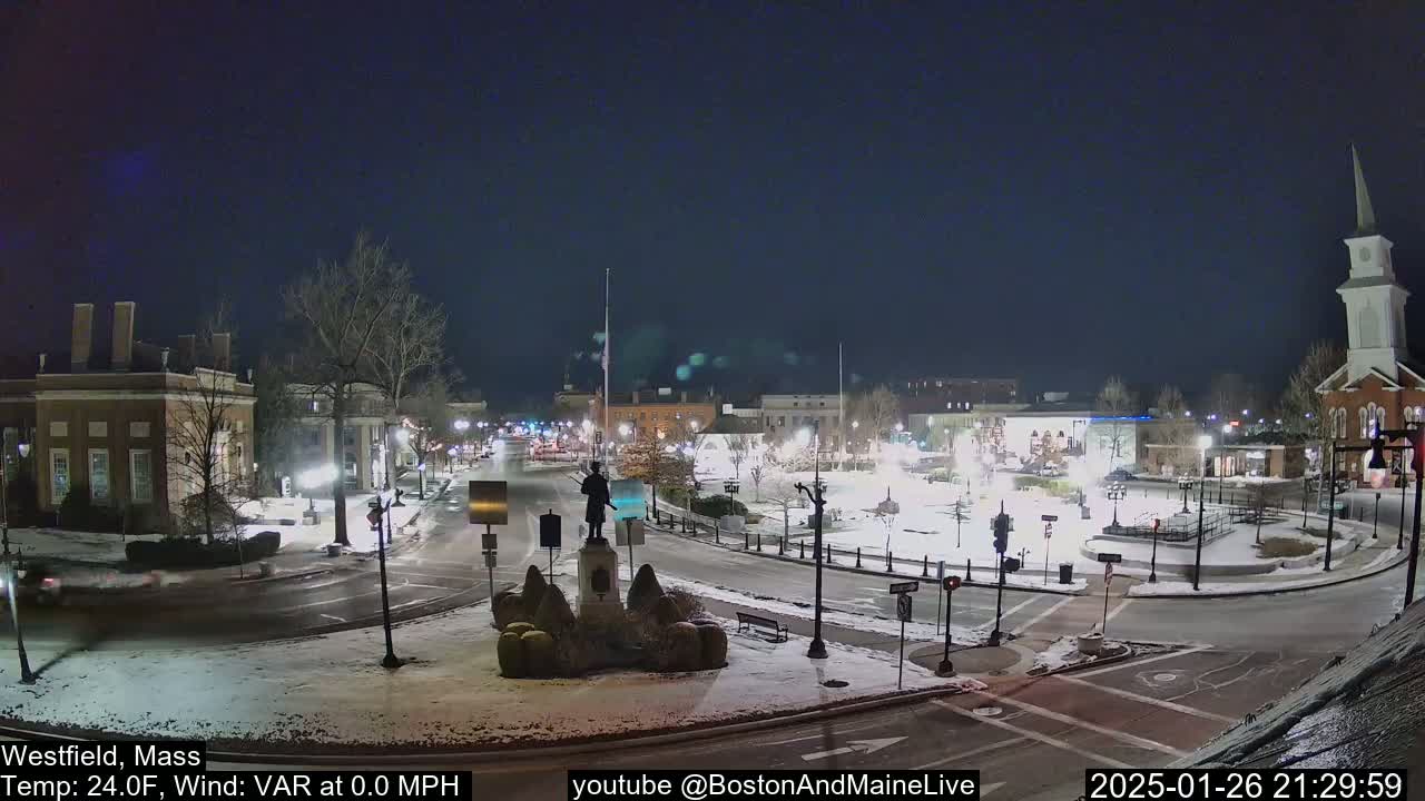 A snow-covered town square at night is illuminated by streetlights, featuring a statue in the center and buildings surrounding the square.