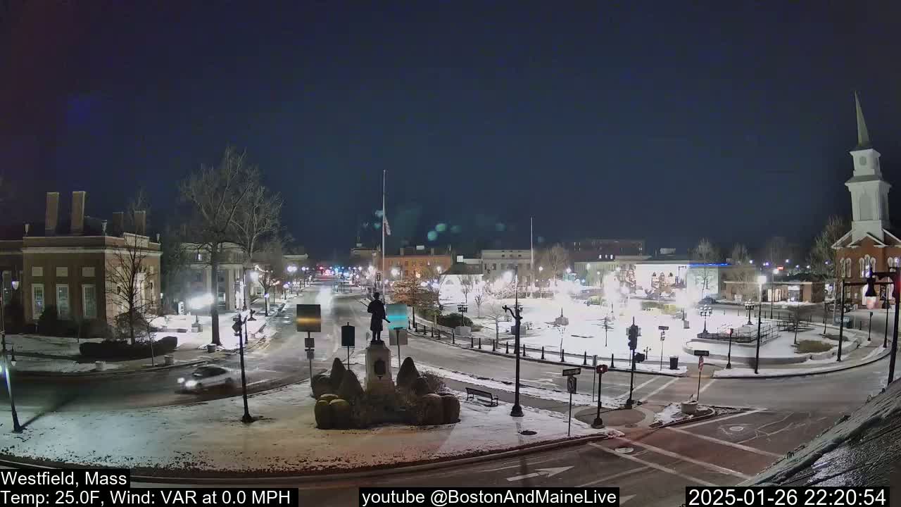 A snow-covered town square at night, illuminated by streetlights, with a statue in the center and several buildings surrounding it.