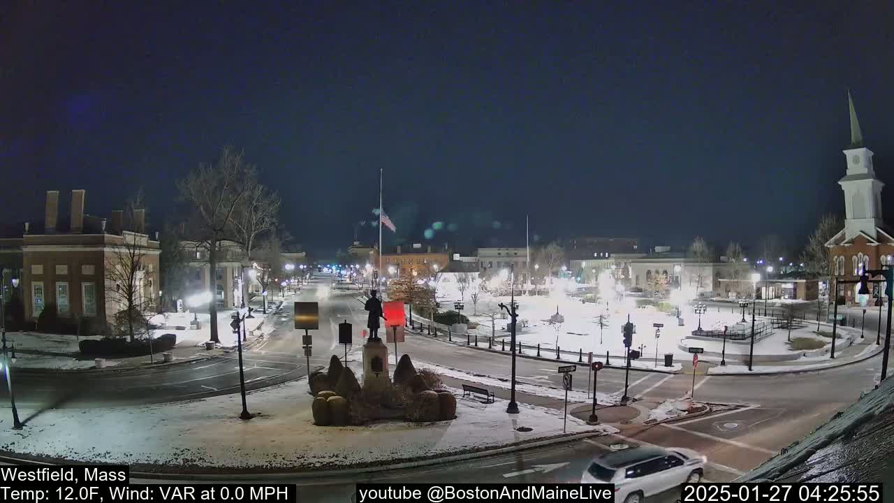 A snow-covered town square at night, illuminated by streetlights, features a monument in the center and several buildings surrounding it.
