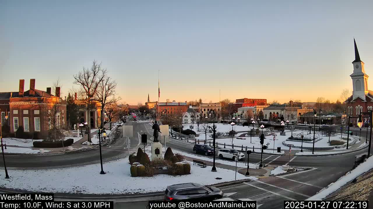 Here is a description of the image in a single sentence: A snow-covered town square with brick buildings, a statue, and cars, is bathed in the soft light of a sunrise under a clear sky.