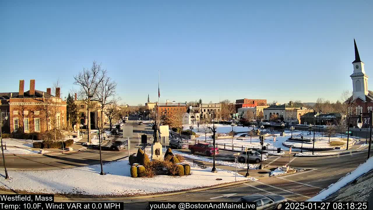 A snow-dusted town square with brick buildings, a monument, and a church steeple under a clear blue sky.
