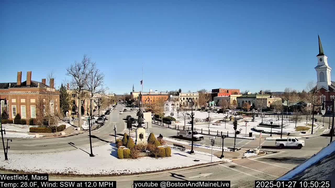 A snow-dusted town square with brick buildings, a church steeple, and vehicles, under a clear blue sky.