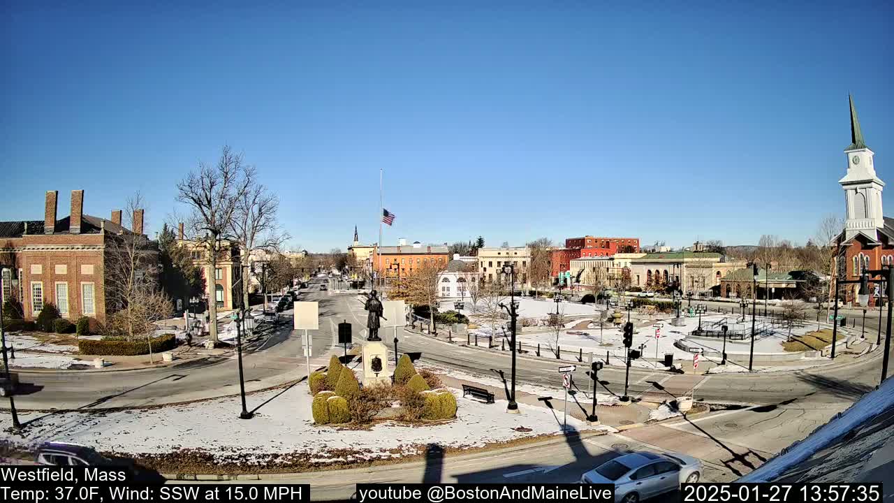 A town square with brick buildings, a statue, and a church steeple under a clear blue sky, with patches of snow on the ground.