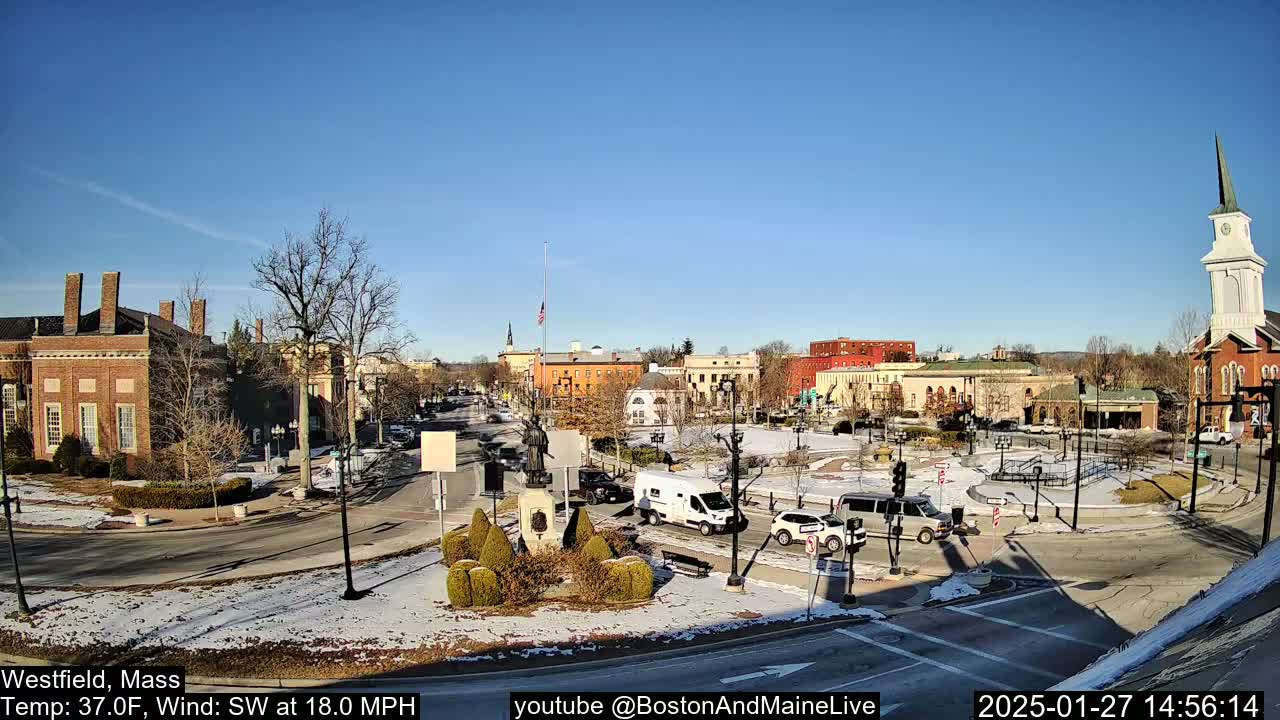 A town square with brick buildings, a monument, and sparse traffic is partially covered in snow under a clear blue sky.