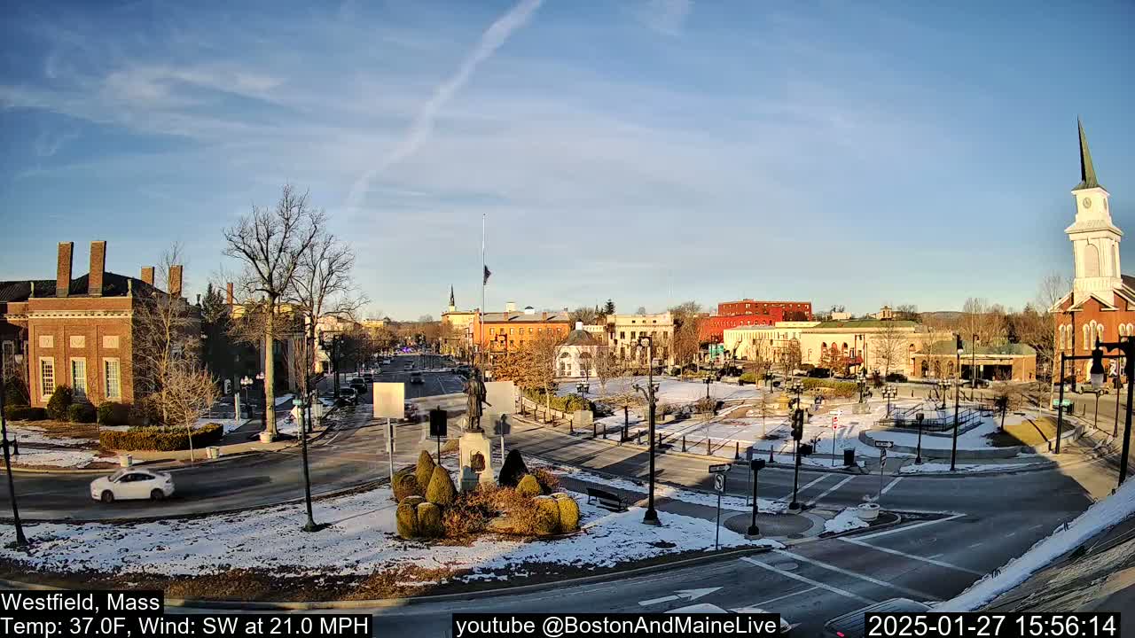 A partly sunny, snowy town square with brick buildings, a monument, and a church steeple under a southwest wind.