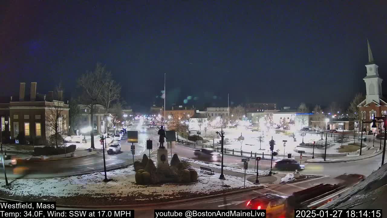 A snow-covered town square at night with a few cars driving around a central monument, under a dark, clear sky.