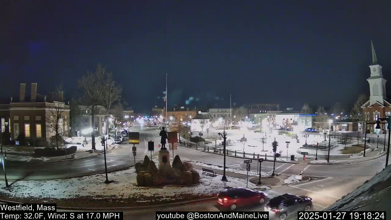 A snow-covered town square at night, with a statue in the center, is illuminated by streetlights under a dark, clear sky, and a few cars are visible on the roads.