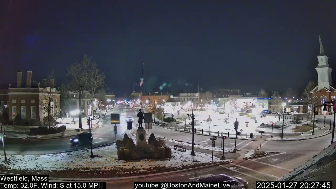 A snow-covered town square at night with a statue in the center,  lighted buildings surrounding it, and a few cars driving on the roads.