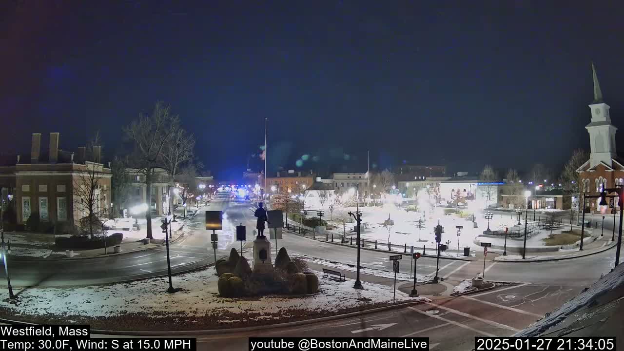 A nighttime view of a town square, lightly snow-covered, with a statue in the center, under a clear, dark sky, with a moderate south wind.