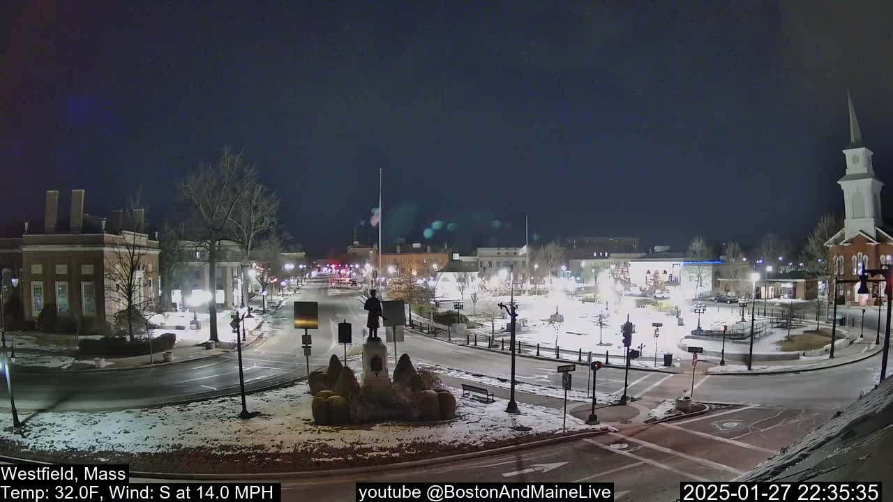 A nighttime view of a town square with snow on the ground, under a dark sky, featuring a statue in the center and buildings surrounding it.