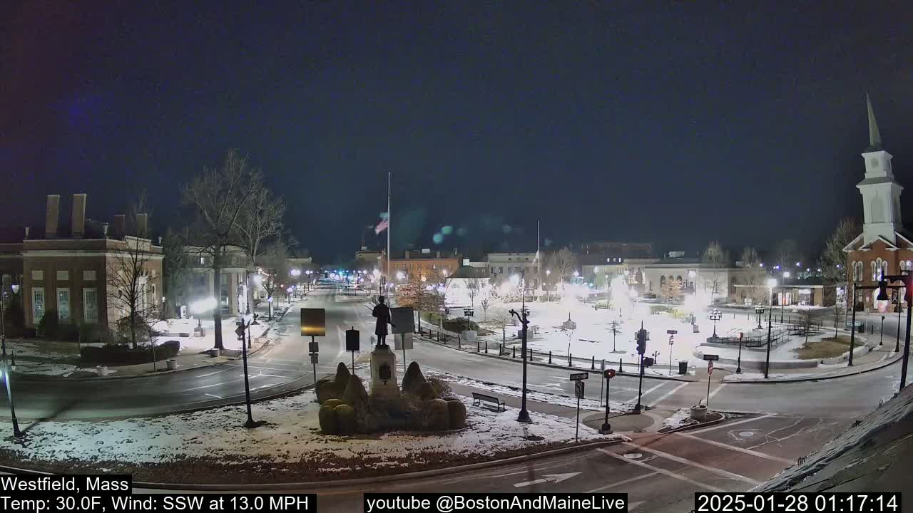 A snow-covered town square at night, illuminated by streetlights, features a statue in the center and several buildings surrounding it, with a light wind blowing from the southwest.