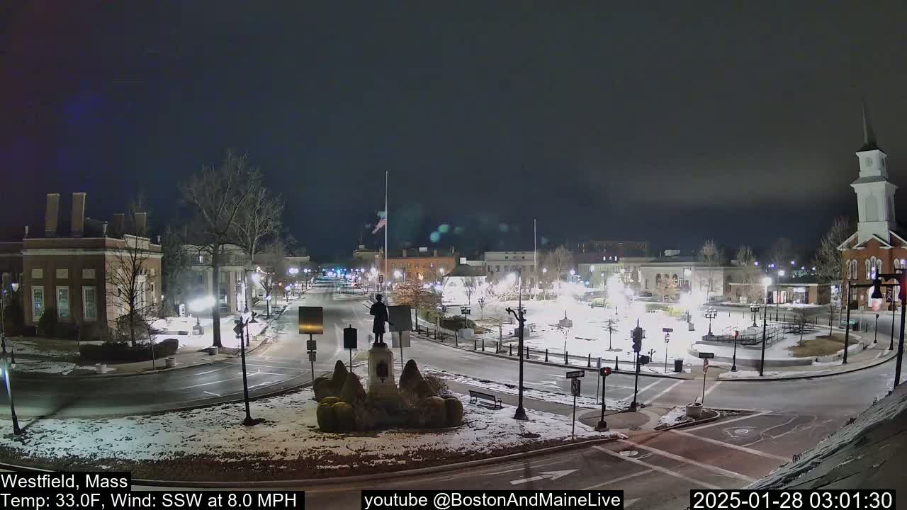 A nighttime view of a snow-covered town square with a statue in the center, under a dark sky.