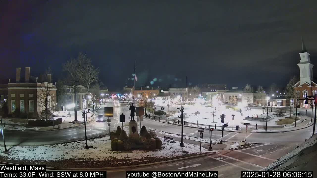 A snow-covered town square at night, illuminated by streetlights, with a statue in the center and several buildings surrounding it.