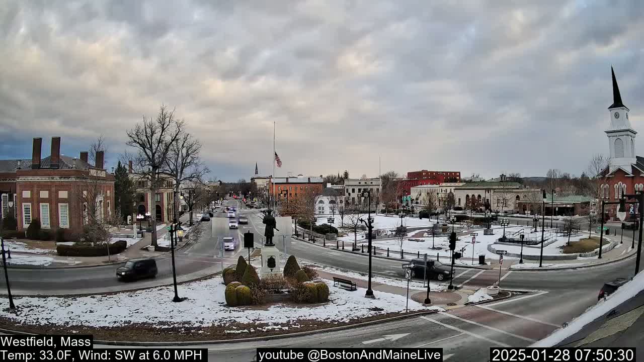 A snow-dusted town square with brick buildings, a statue, and sparse traffic under a cloudy sky.