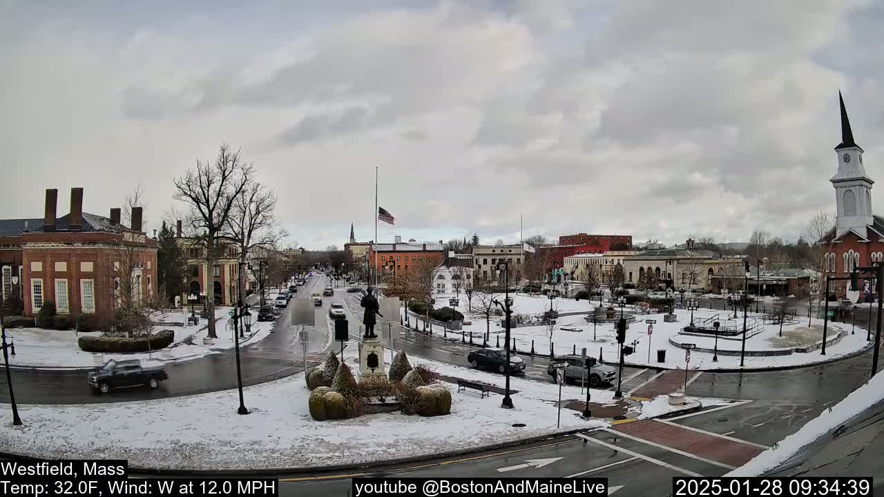 A snow-covered town square with several brick buildings, a statue, and cars driving on wet roads under a cloudy sky.