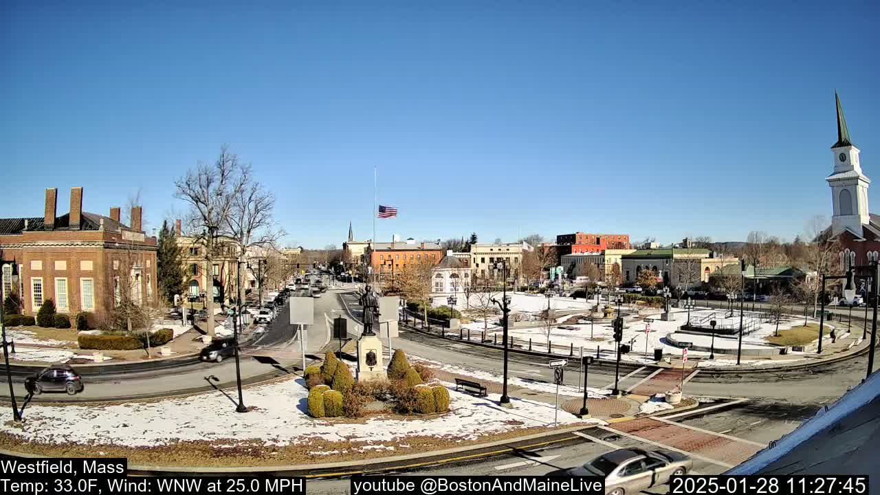 A partly snowy town square with brick buildings and a church steeple under a clear blue sky.