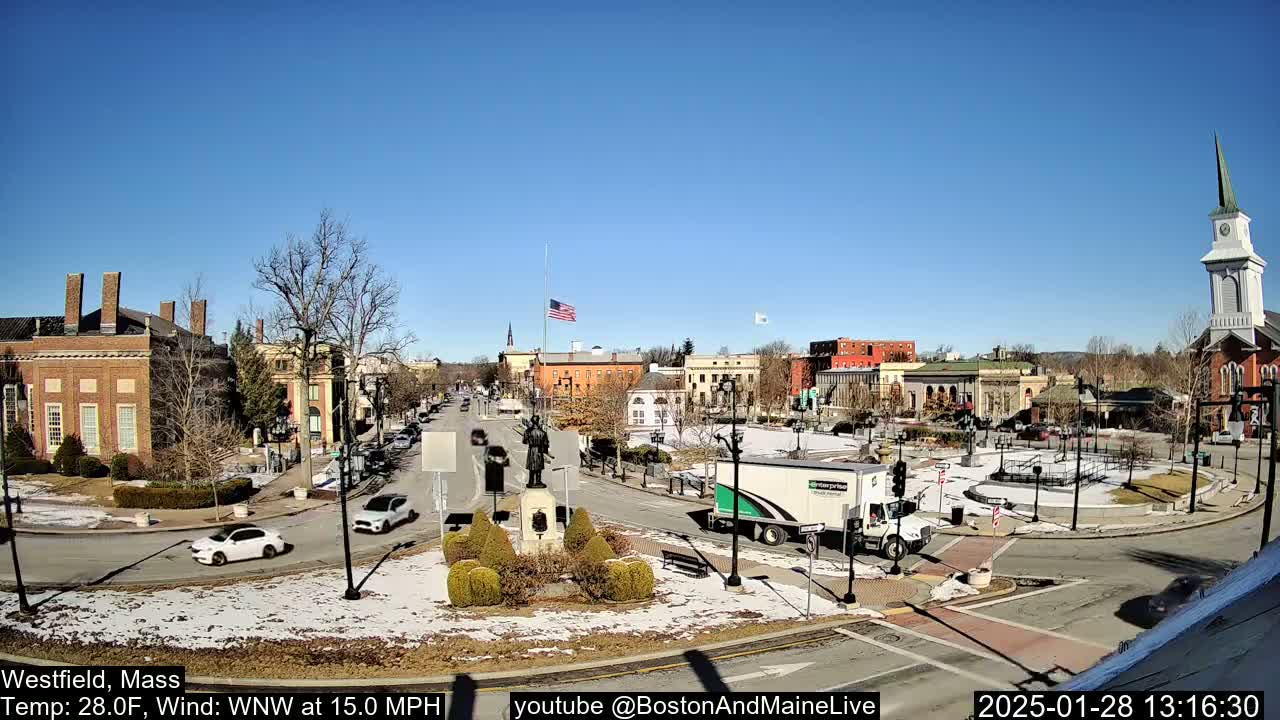 A town square with several buildings, a statue, and sparse traffic on a sunny, cold day with some snow on the ground.