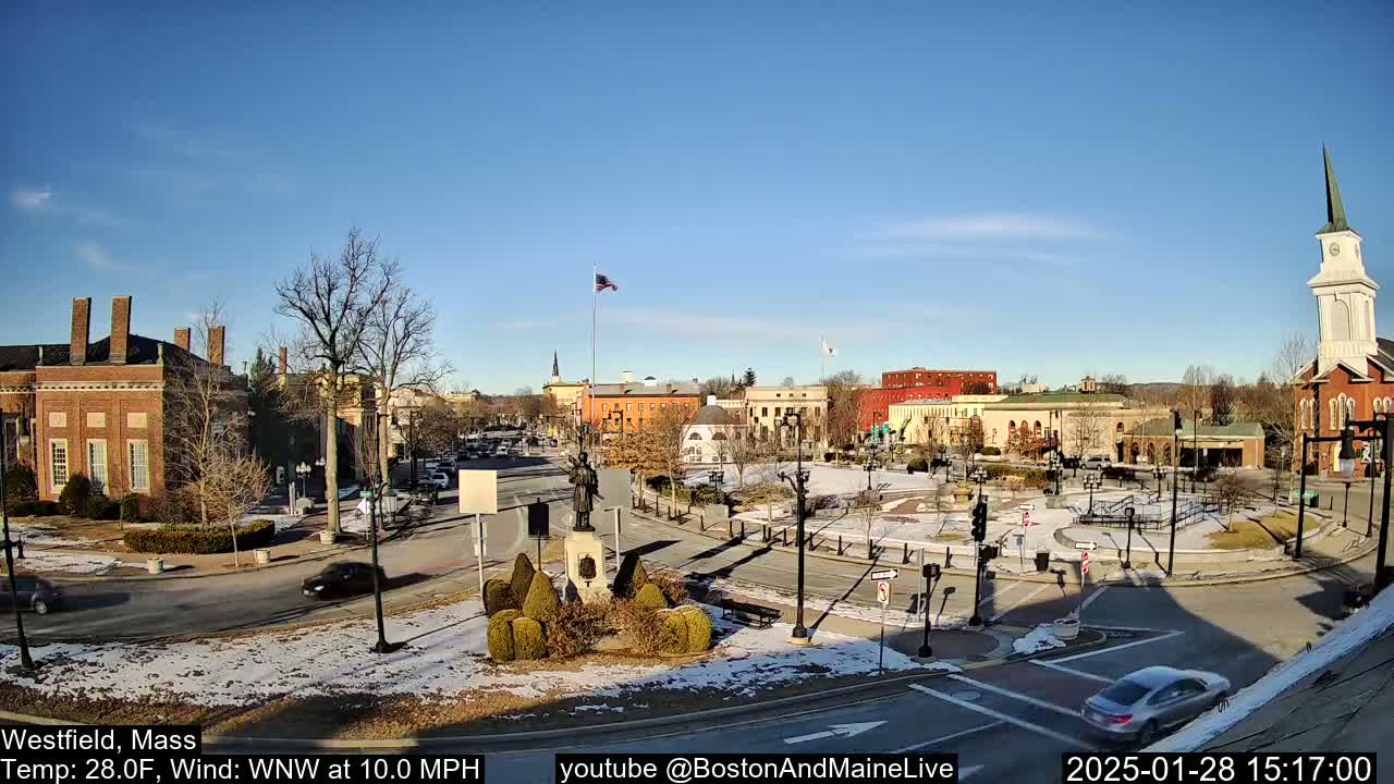 A sunny, cold day in a town square with brick buildings, a statue, and a church steeple, with some snow on the ground and cars driving around.