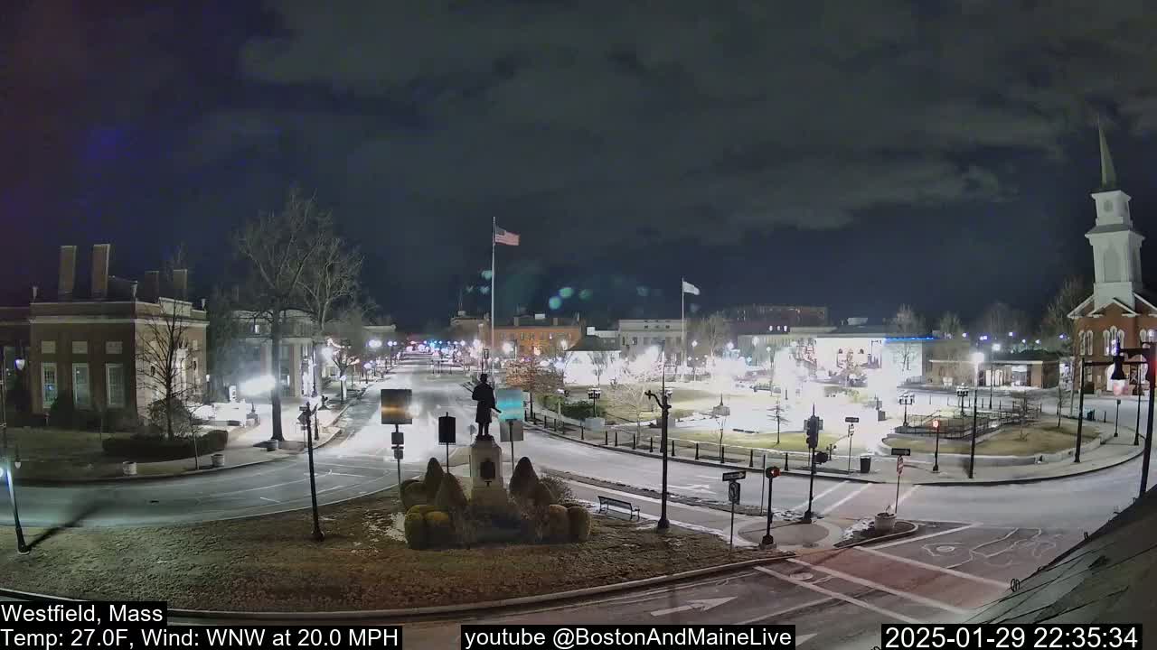 A nighttime view of a town square with light snow on the ground, under a dark, cloudy sky, with a statue in the center and a few cars visible on the roads.