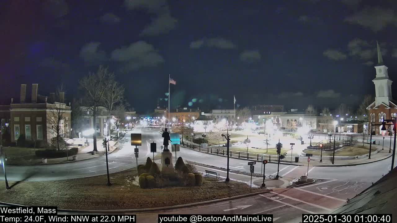 A nighttime view of a town square, with a statue in the center, under a partly cloudy sky and a strong northwest wind.