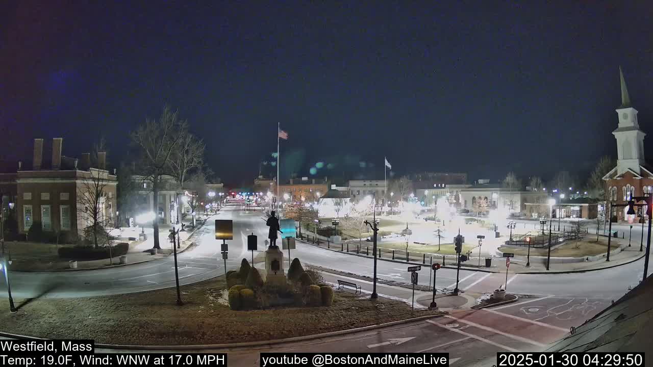 A nighttime view of a town square with a statue at its center, under a clear, dark sky, with a light wind.