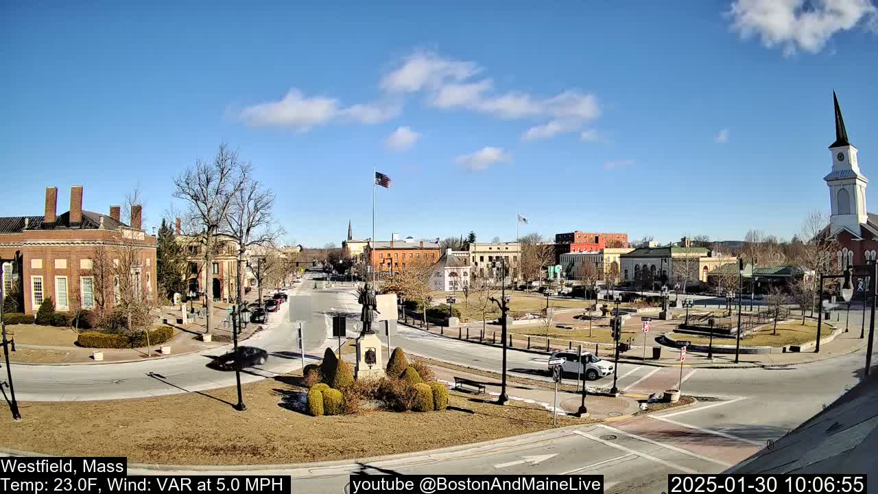 A sunny day reveals a town square with brick buildings, a statue, and a church steeple, surrounded by a sparsely-leafed landscape.