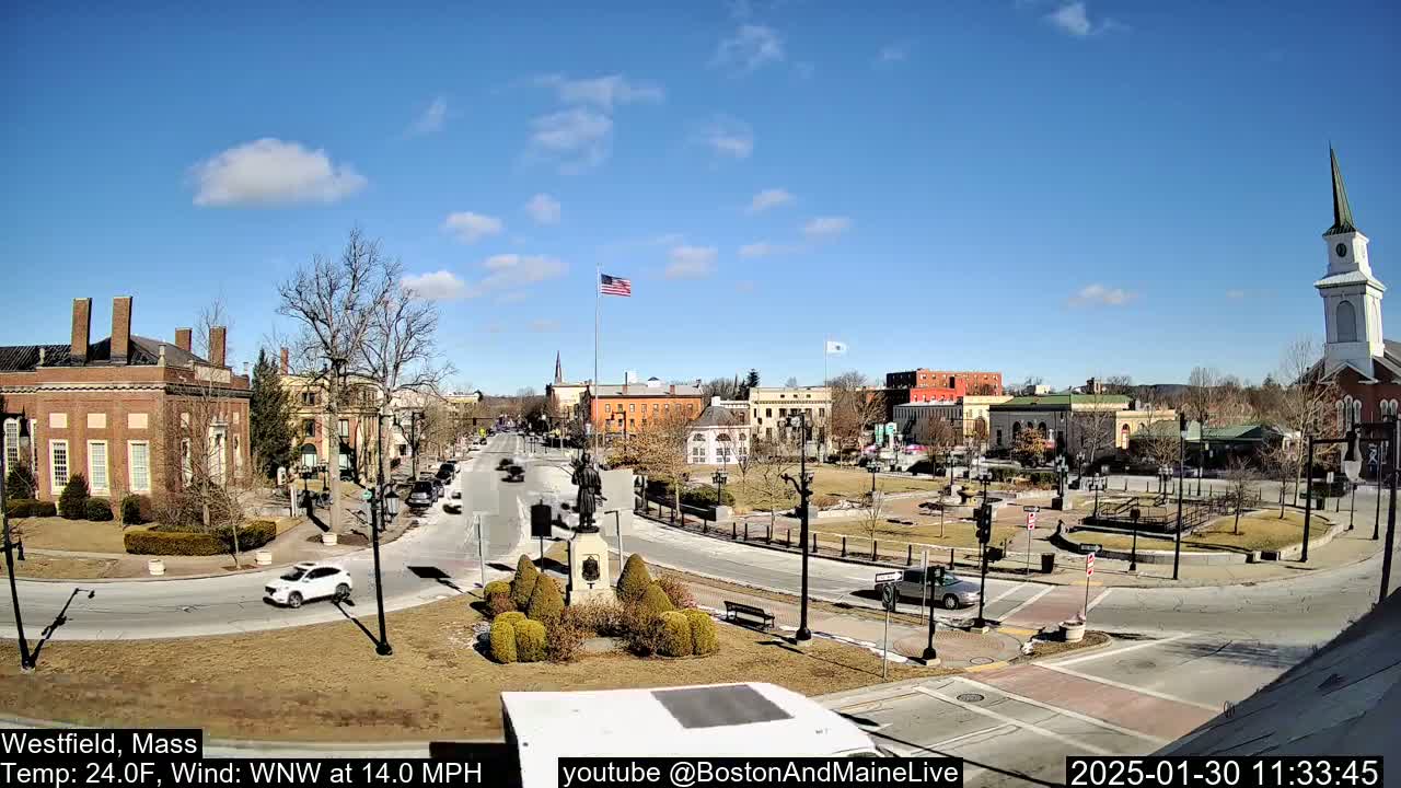 A sunny day reveals a town square with brick buildings, a statue, and sparse traffic under a mostly clear blue sky.