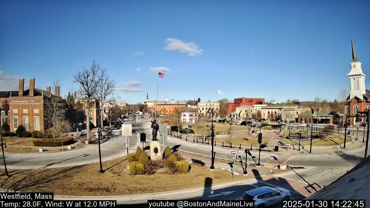 A town square with brick buildings, a monument, and a church steeple under a mostly sunny sky.