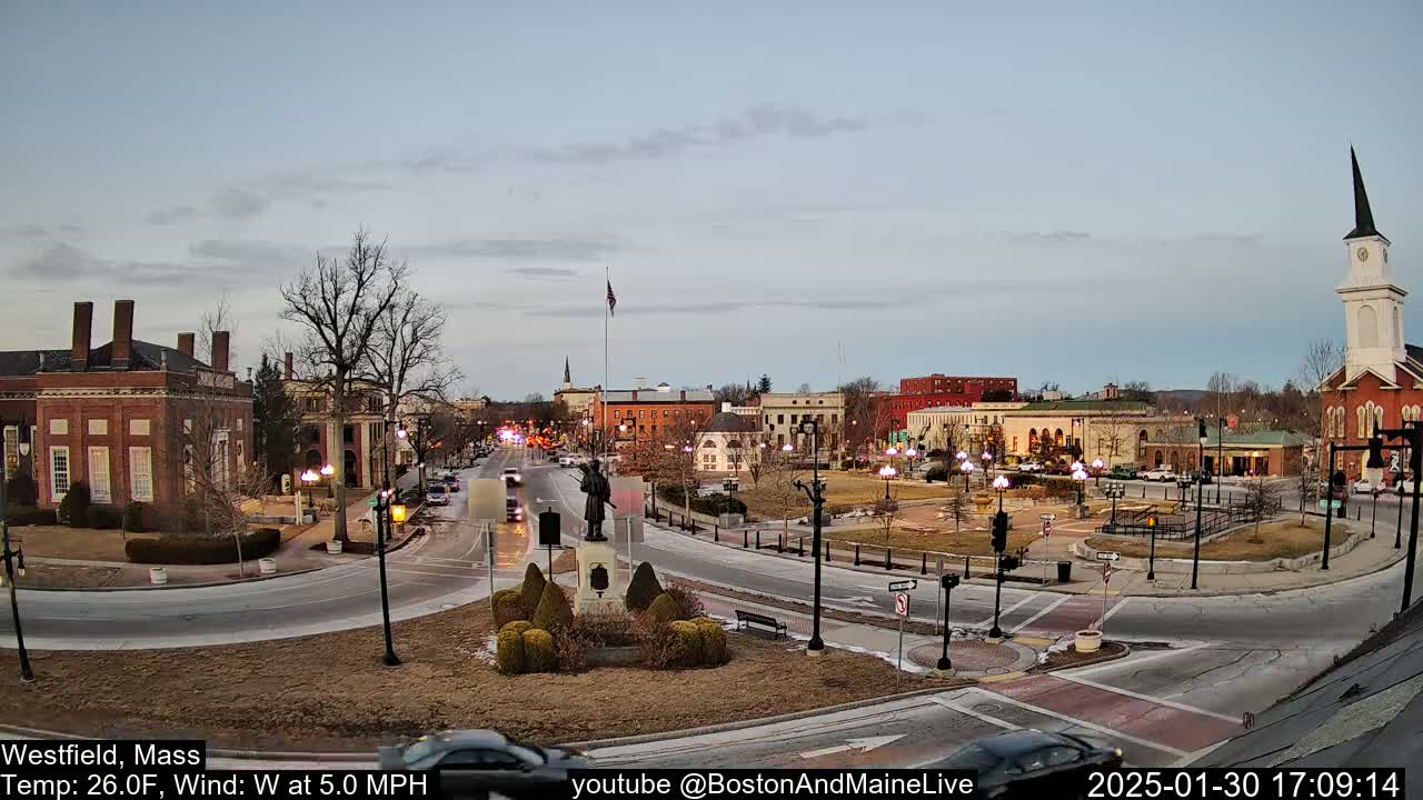 A town square with brick buildings, a statue, and cars, under a partly cloudy twilight sky.