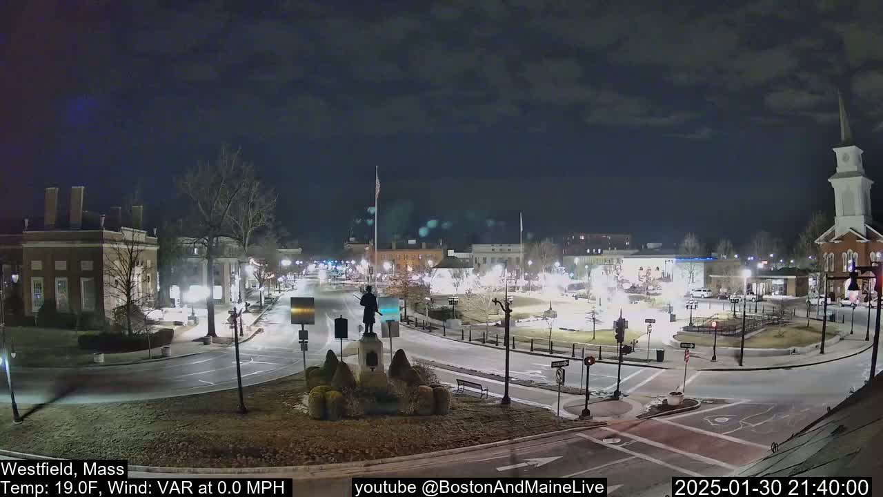 A nighttime view of a town square with a statue at its center, surrounded by buildings and sparse traffic under a cloudy sky.