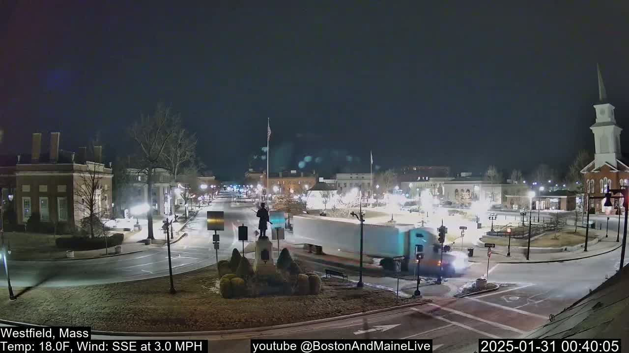 A nighttime view of a town square with a statue in the center, lightly snow-covered ground, and a large truck driving through the intersection under a dark, clear sky.