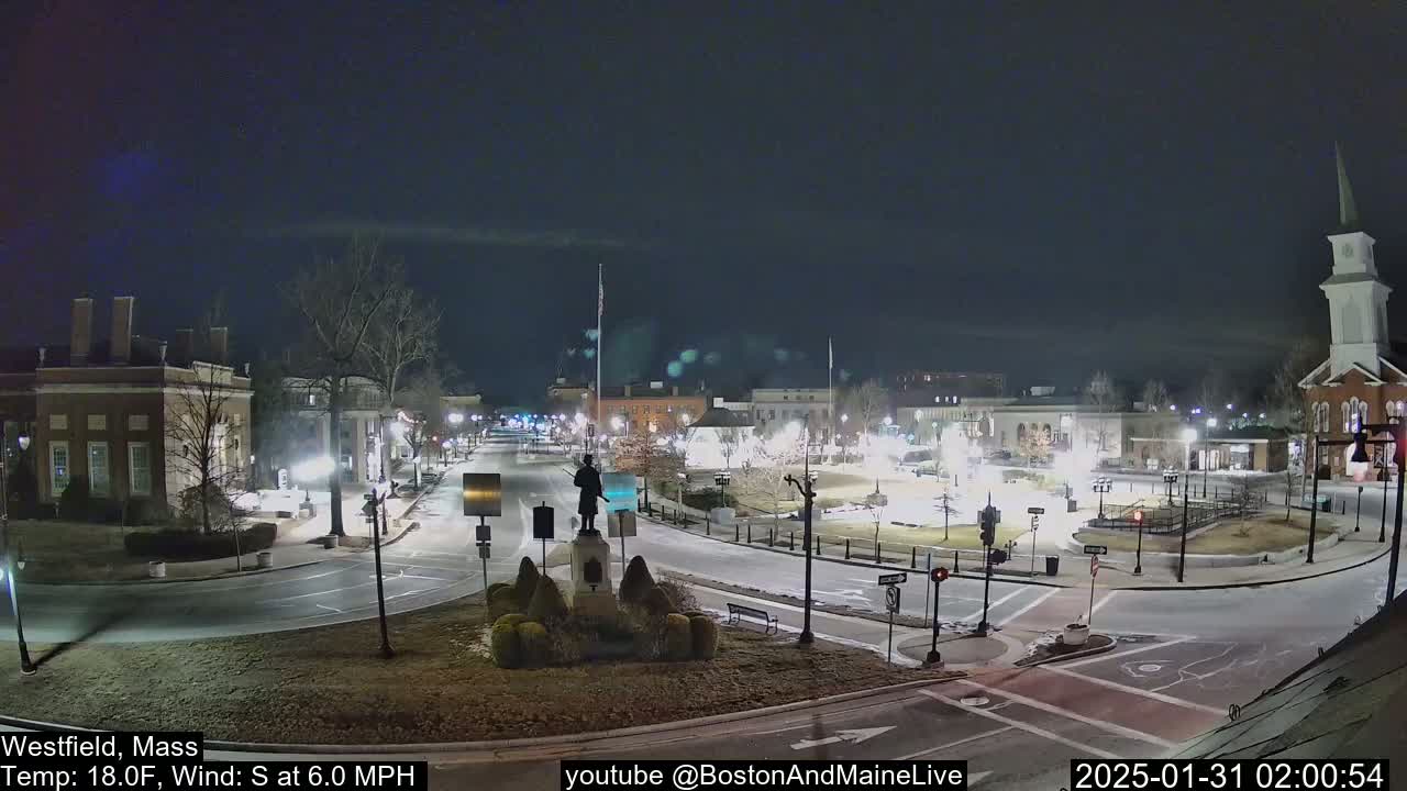 A nighttime, mostly clear view of a town square with a statue in the center, surrounded by buildings and roads with minimal traffic.