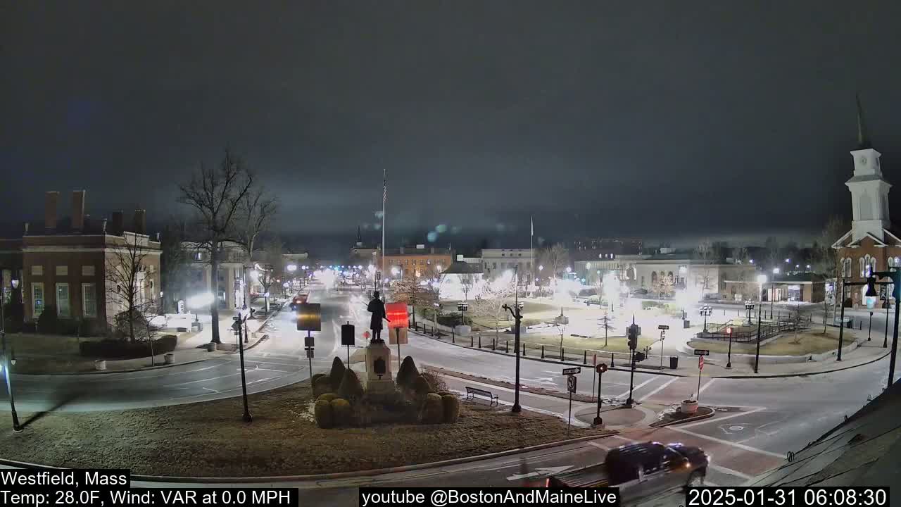 A nighttime view of a town square with a statue in the center, under a cloudy sky with calm winds and a temperature of 28 degrees Fahrenheit.