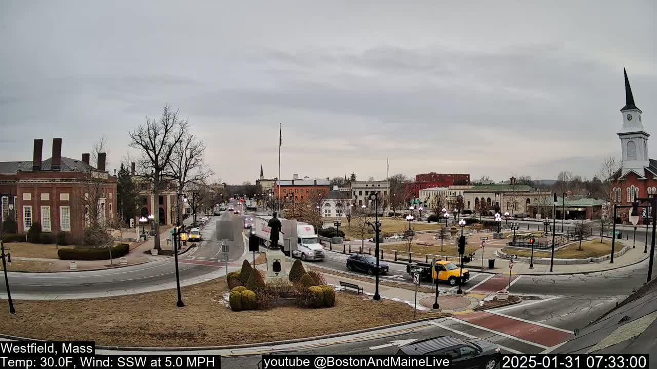 A town square with brick buildings, sparse trees, and a few cars is visible under an overcast sky.