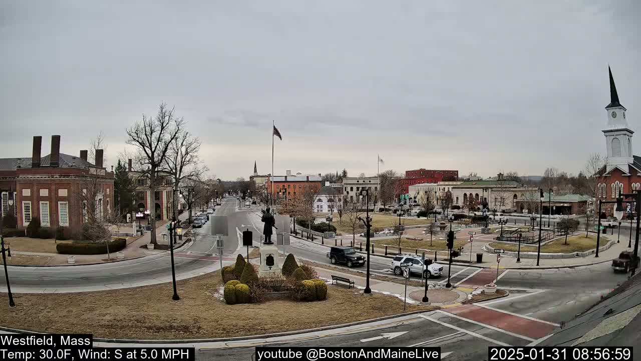 A town square with brick buildings, a statue, and sparse traffic under an overcast sky.