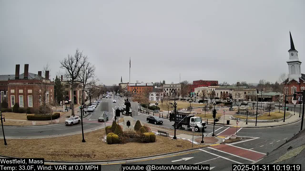 A town square with brick buildings, a church steeple, and sparse traffic under an overcast sky.