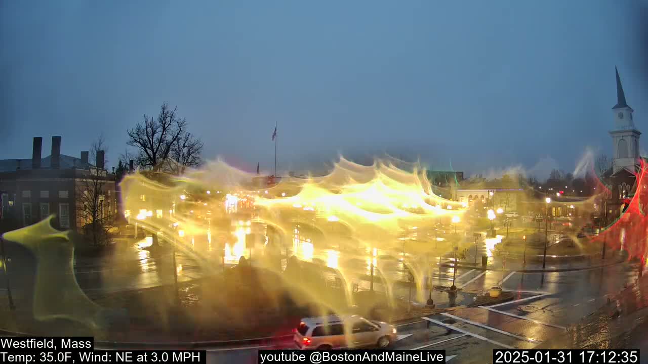 A wet, nighttime town square with buildings, streetlights, and vehicles is illuminated by streaks of yellow light in a 35°F temperature with a northeast wind.