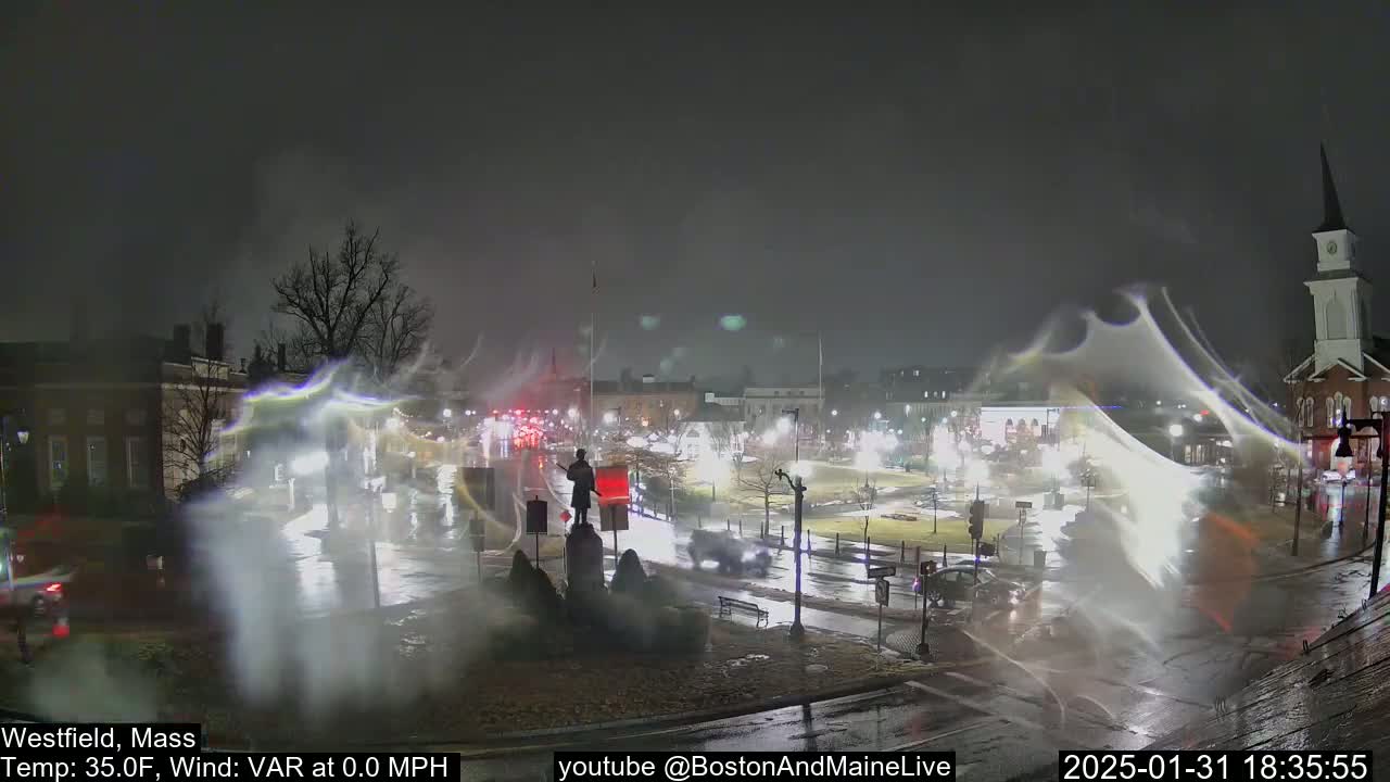 A nighttime view of a town square with a statue in the center, wet streets, and light snow or blowing debris in the air.