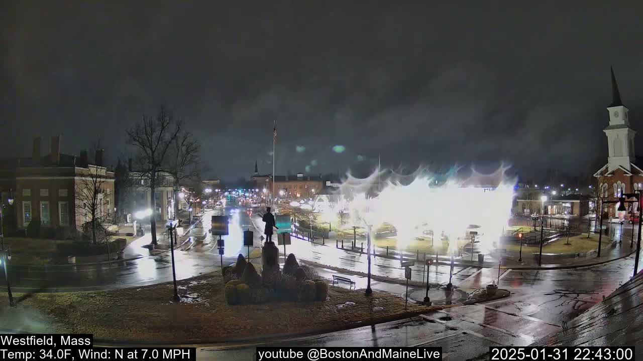 A nighttime view of a town square with wet roads and a statue in the center, under a dark sky with bright, blurry lights in the background.